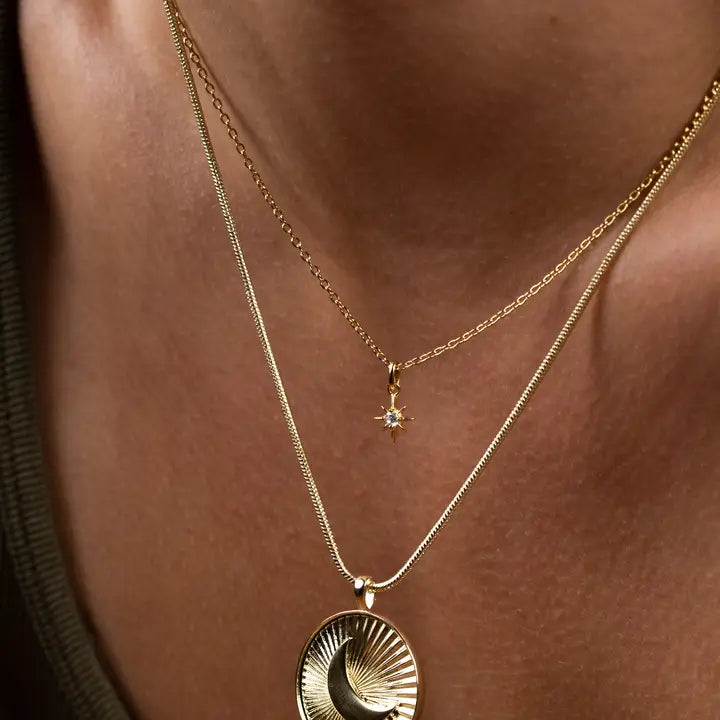 Close-up of two gold necklaces: JaxKelly Tiny Star Necklace, 18k gold plated with a star-shaped Cubic Zirconia pendant, and another featuring a larger round textured, swirling pendant on an adjustable chain.