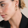 A woman with light skin and freckles wears JaxKelly's Puffed Gold Heart Earrings—vintage-inspired studs with 18K gold plating—in her left ear, alongside a gold hoop earring. She has her hair pulled back and is wearing a black top.