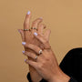 Two hands with light purple nail polish and multiple gold rings, including the JaxKelly White Opal Inset Baguette, are shown against a beige background. The person wears a dark long-sleeve garment.