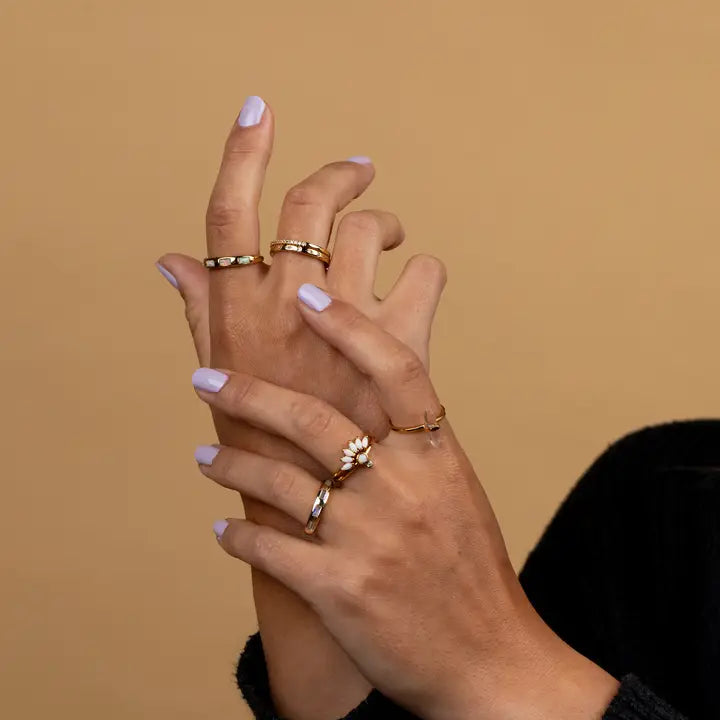 Two hands with light purple nail polish and multiple gold rings, including the JaxKelly White Opal Inset Baguette, are shown against a beige background. The person wears a dark long-sleeve garment.