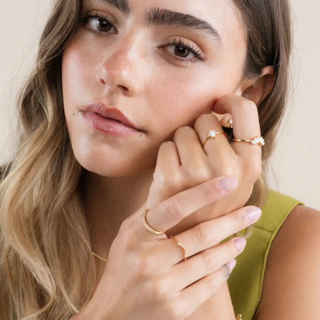 A woman with wavy light brown hair in a green top poses with her hands near her face, showcasing the Opal Crown Ring by JaxKelly along with gold jewelry and a delicate necklace. She has natural makeup and a calm expression.
