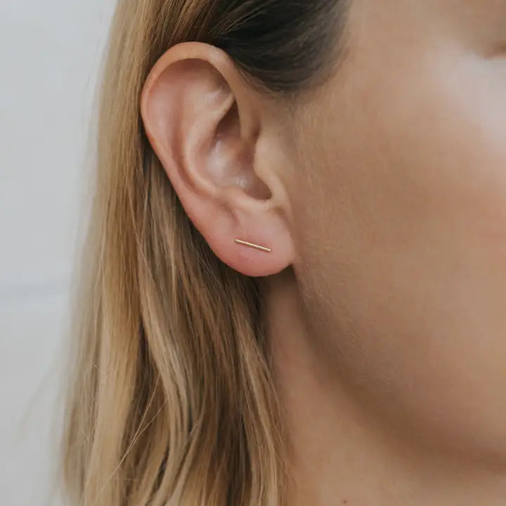 Close-up of a person's ear with straight, light brown hair wearing the JaxKelly Minimalist Bar earrings. These tarnish-free gold studs highlight smooth skin against a softly blurred background.