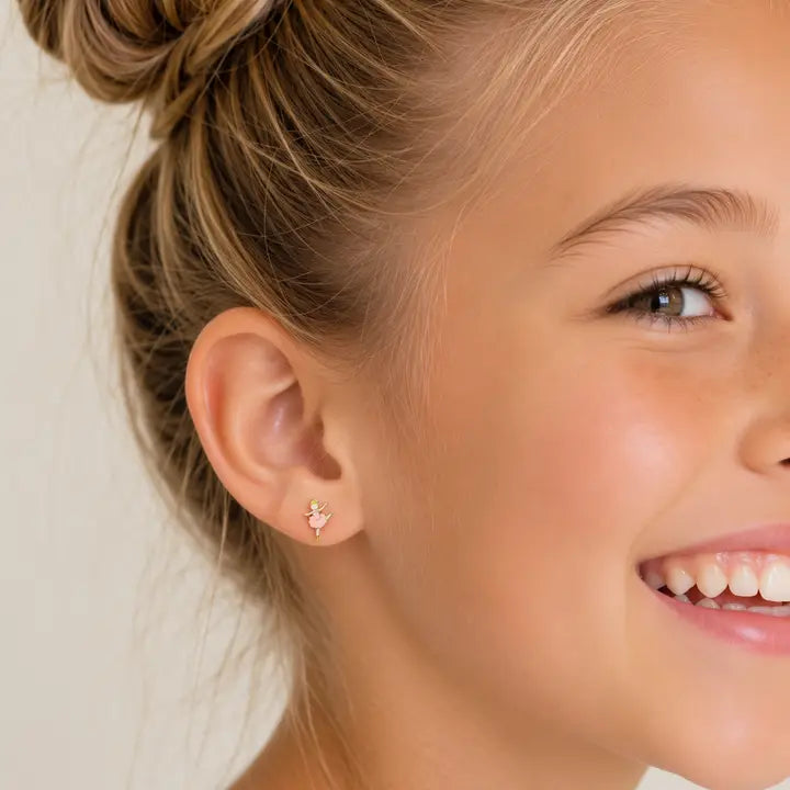 A young girl wears hypoallergenic JaxKelly Ballerina Enamel Flat Back Kids Earrings shaped like small gold ballerinas in pink tutus.