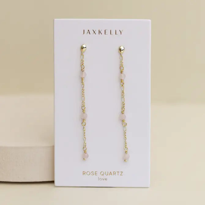 A pair of JaxKelly Gemstone Chain Drop - Rose Quartz earrings with 18k gold plating and rose quartz beads, shown on a white card labeled "JaxKelly" and "Rose Quartz love" against a neutral background.