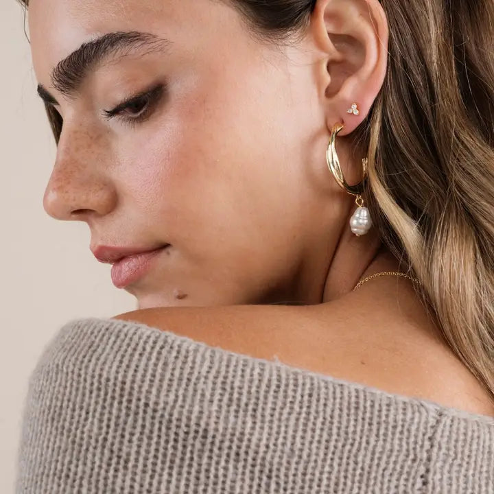 A woman with light skin and freckles wears JaxKelly's Twisted Pearl Drop Hoop - Gold Earrings, featuring gold-plated hoops and a dangling pearl. She pairs them with a small stud earring and a beige off-shoulder knit sweater against a neutral background.