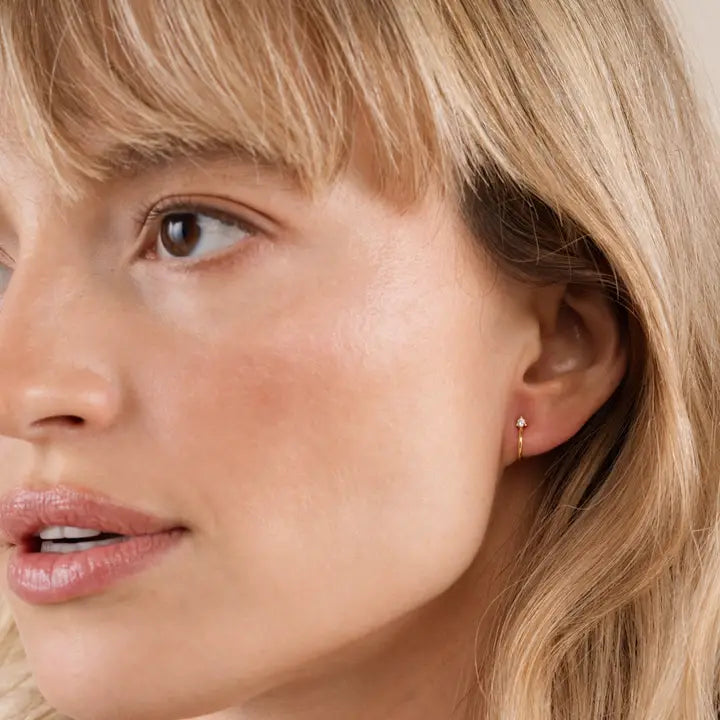 A close-up of a blonde woman wearing JaxKelly's Open Mini Hoop Earring - White, featuring a small dangling charm. She has smooth, natural makeup and a neutral expression.