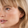 A close-up of a blonde woman wearing JaxKelly's Open Mini Hoop Earring - White, featuring a small dangling charm. She has smooth, natural makeup and a neutral expression.