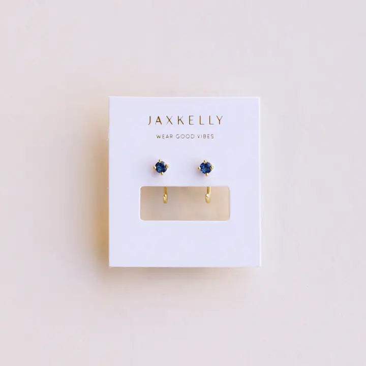 A pair of JaxKelly Sapphire CZ Huggies with dark blue stones is displayed on a white card reading "JAX KELLY WEAR GOOD VIBES" against a light background.