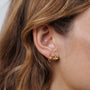 A close-up shows a person with light brown wavy hair wearing JaxKelly's Petite Gold Knot Earrings, crafted with 18K gold plating. The neutral, blurred background highlights the elegant studs—perfect for everyday wear.