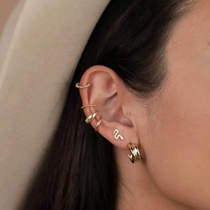 A close-up of a woman with dark hair in a beige hat showcases her unique style, wearing JaxKelly’s Bold Hoop gold earrings paired with a small squiggle stud on sterling silver posts.