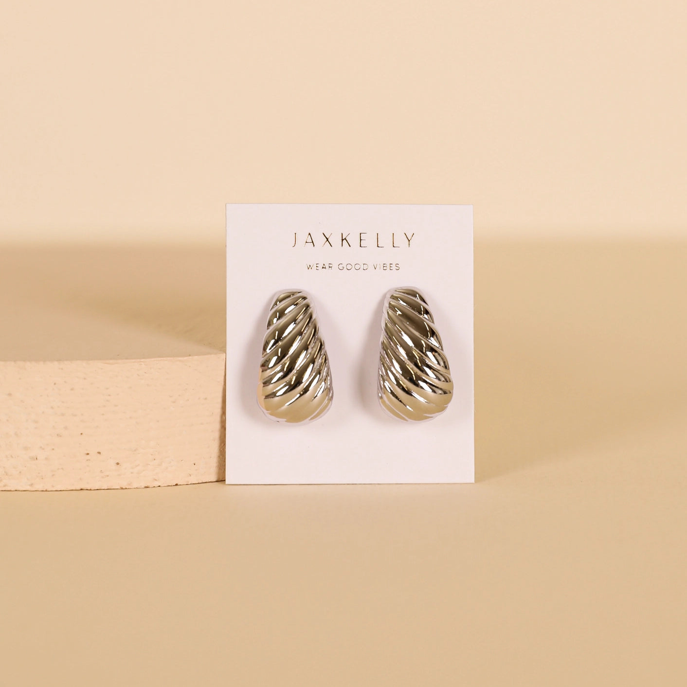 The Silver Bold Swirl - Earring is shown as a pair of sterling silver, ridged hoops on a white JaxKelly card with a beige background. These earrings feature a rounded, textured swirl design.