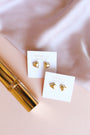 Puffed Gold Heart Earrings with 18K gold plating are displayed on white JaxKelly cards atop a light pink surface, next to a gold cylinder and silky light pink fabric.