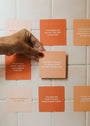 A hand with painted nails places a Positivity Shower Affirmations™ reusable card on a tiled wall, surrounded by other orange and peach cards that display motivational quotes in white text.