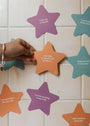 A hand places an orange star-shaped card on white tiles. Kids Shower Affirmations™—colorful, waterproof cards with positive quotes like "I am kind to myself and to others"—are displayed on the wall.