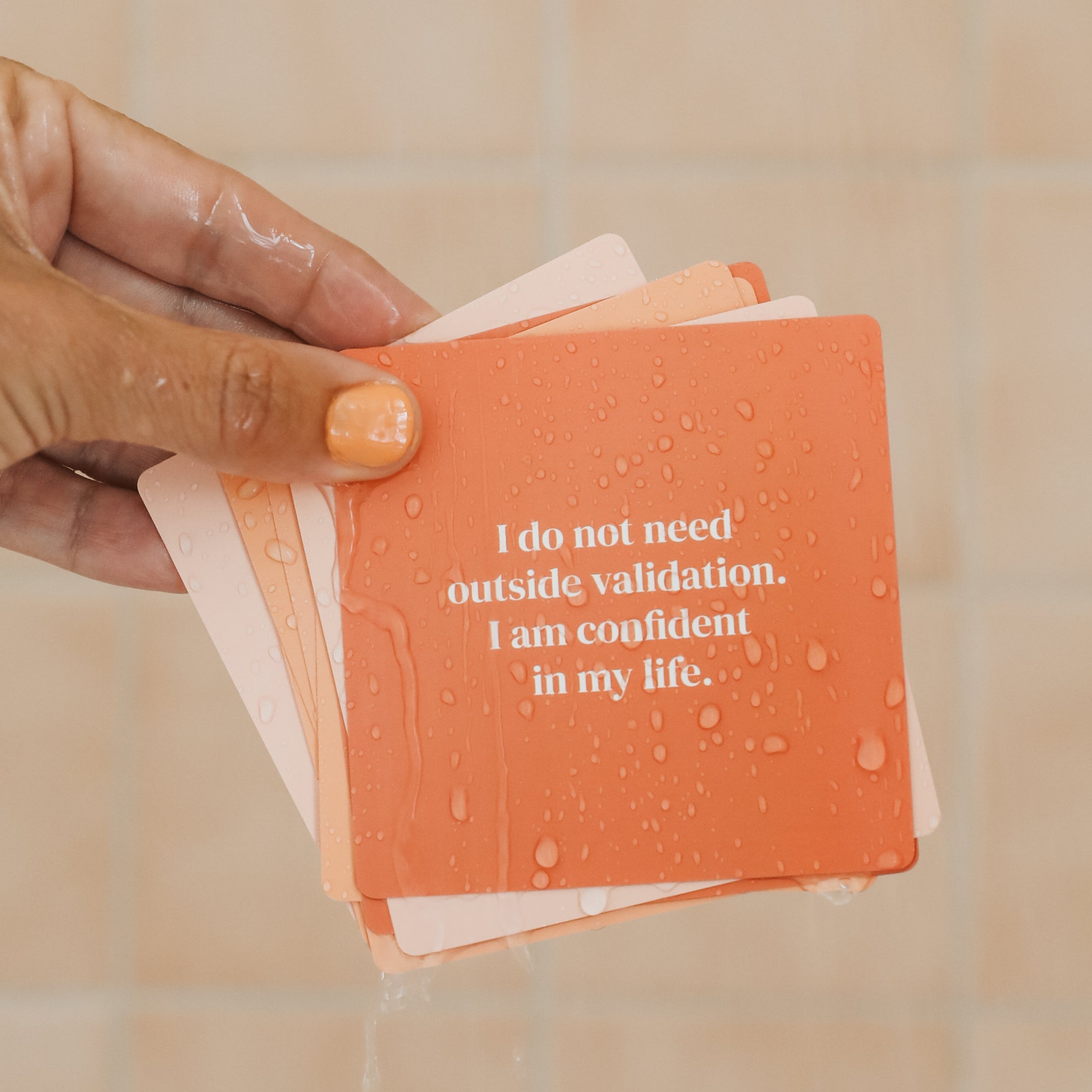 A hand with orange-painted nails holds Positivity Shower Affirmations™, reusable waterproof cards featuring quotes like "I do not need outside validation. I am confident in my life." against a peach-colored tiled wall.