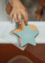 A child’s soapy hand reaches for Kids Shower Affirmations™—pastel, waterproof cards on the bathtub edge, including one that reads, “I am kind to myself and to others.”.