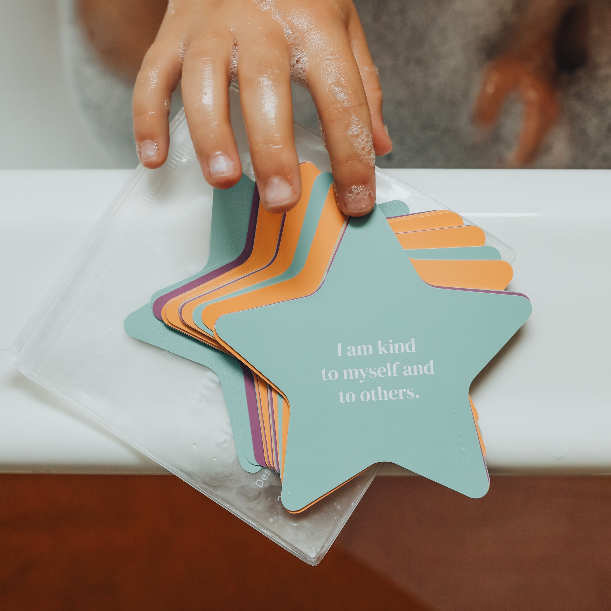A child’s soapy hand reaches for Kids Shower Affirmations™—pastel, waterproof cards on the bathtub edge, including one that reads, “I am kind to myself and to others.”.