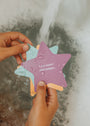 Hands holding colorful Kids Shower Affirmations™ foam star shapes over a bathtub. The purple star says, “I am smart and unique.” Water runs in the background, making these affirmation cards perfect for kids during bath time.