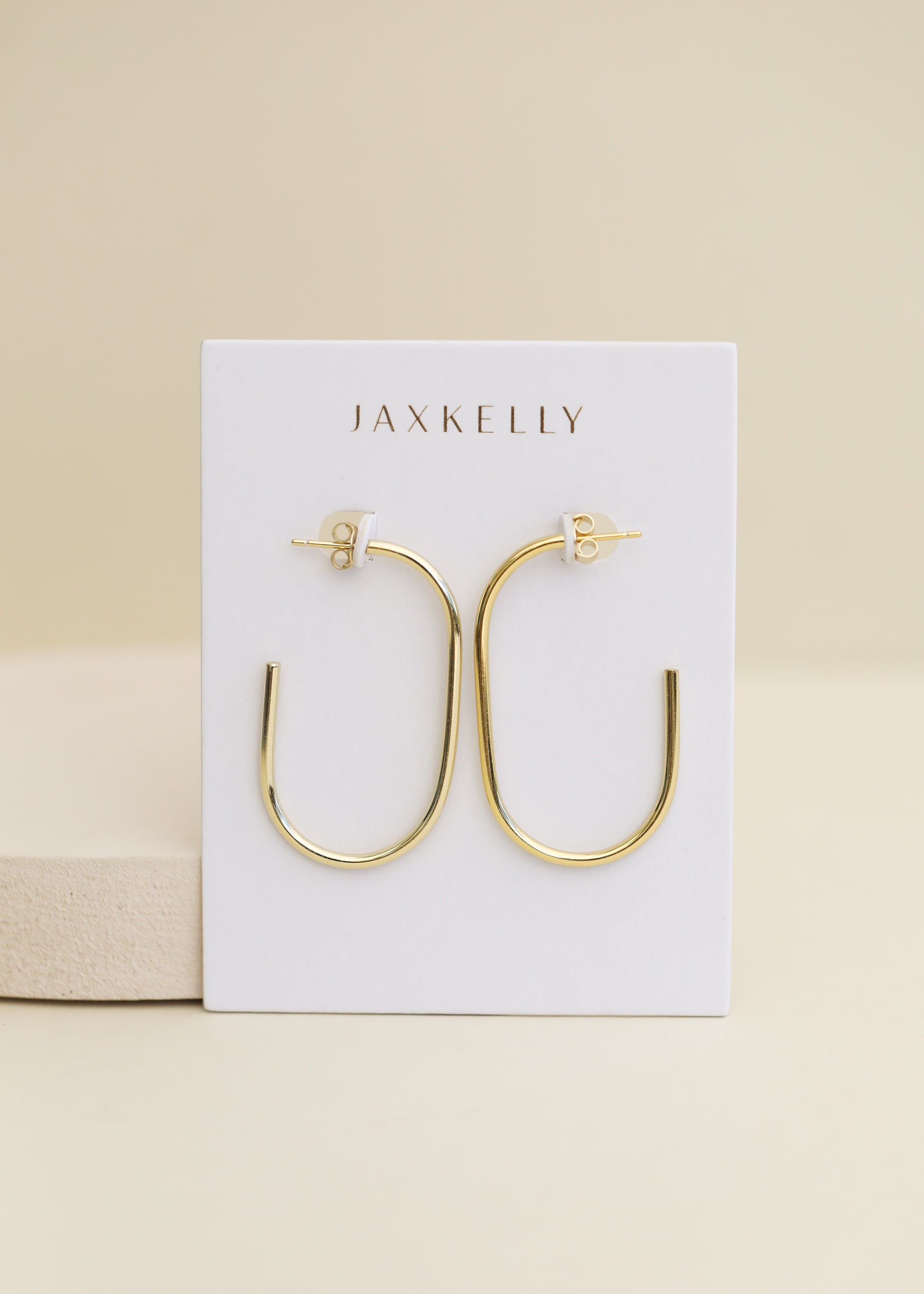 A pair of JaxKelly Gold Large Open Hoop earrings, hypoallergenic and displayed on a white JaxKelly card, set against a neutral beige background.