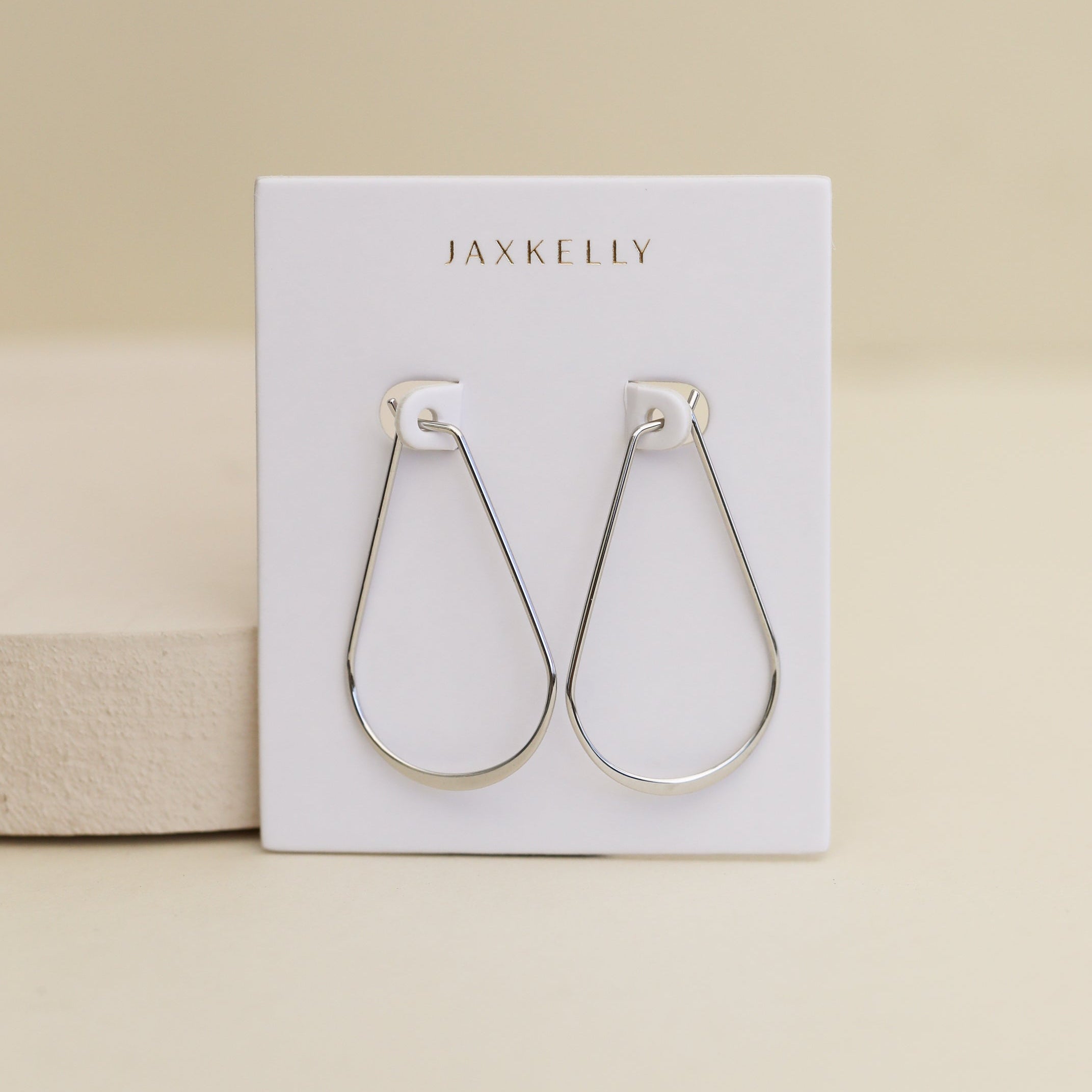 The Silver Long Oval Hoop earrings are minimalist, teardrop-shaped Sterling Silver wire hoops shown on a white JaxKelly branded card against a neutral beige background.