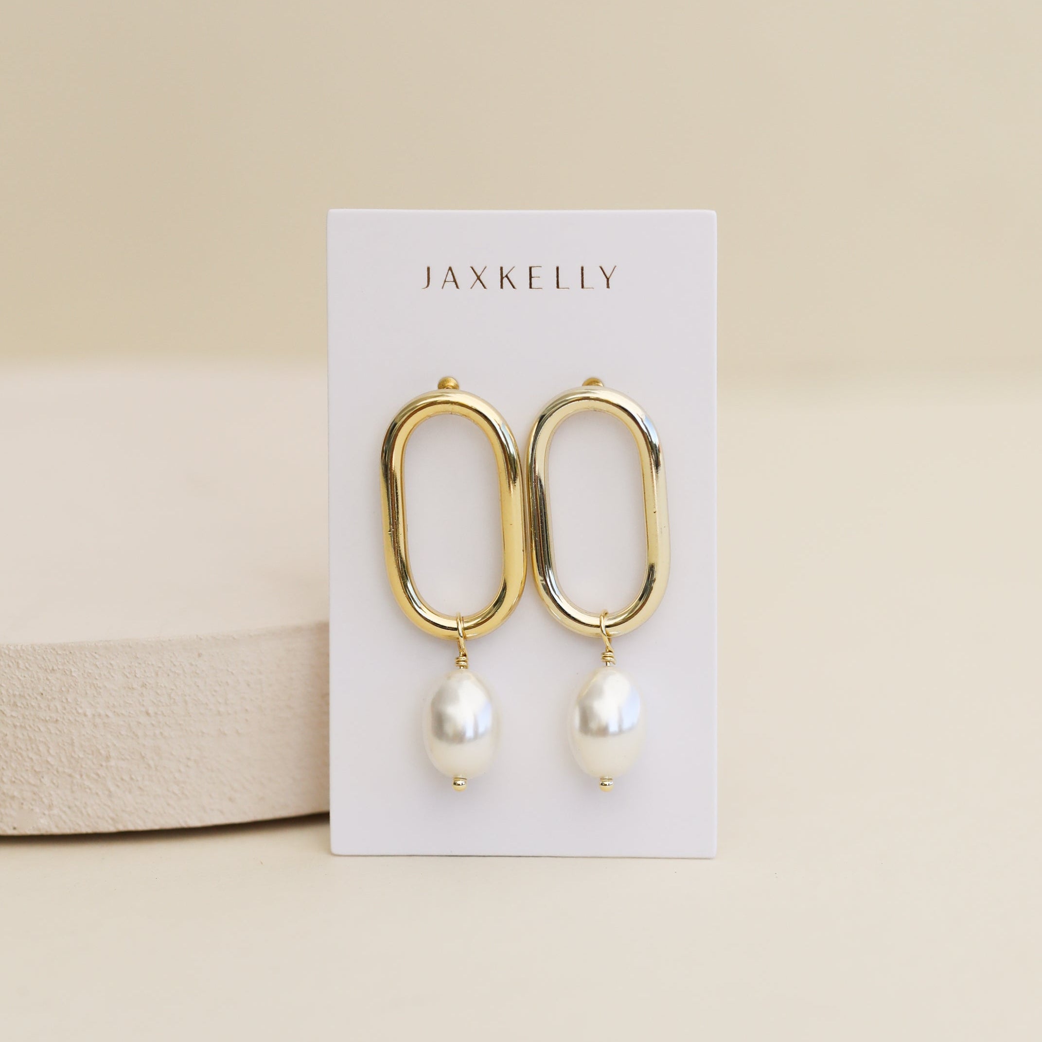 The Oval Pearl Drop earrings feature 18k gold plated elongated hoops with dangling white pearls, showcased on a JaxKelly card against a neutral background.