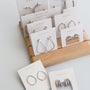 JaxKelly Mystery Earrings are displayed on white cards, neatly arranged on a wooden stand with two extra pairs in front—perfect for fans of surprise jewelry.