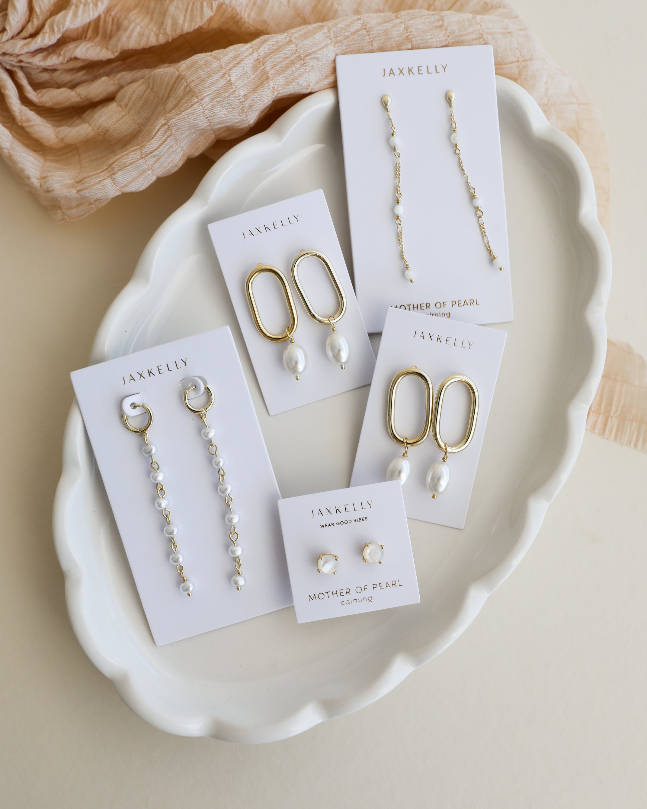Five pairs of pearl earrings on JaxKelly cards arranged on a white ceramic tray, with a beige fabric nearby. The earrings have gold or silver tones and feature various pearl designs.
