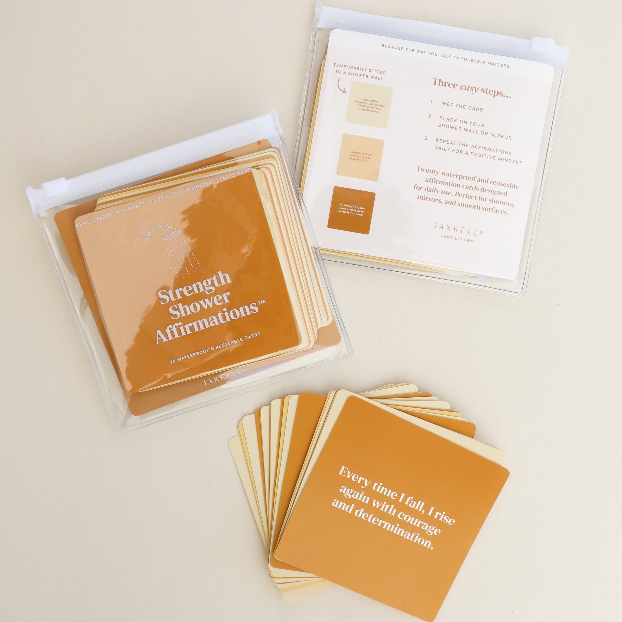 Strength Shower Affirmations™: Square, waterproof affirmation cards in warm brown and beige tones—some stacked, some spread out. The visible card reads, "Every time I fall, I rise again with courage and determination." Reusable quote cards.