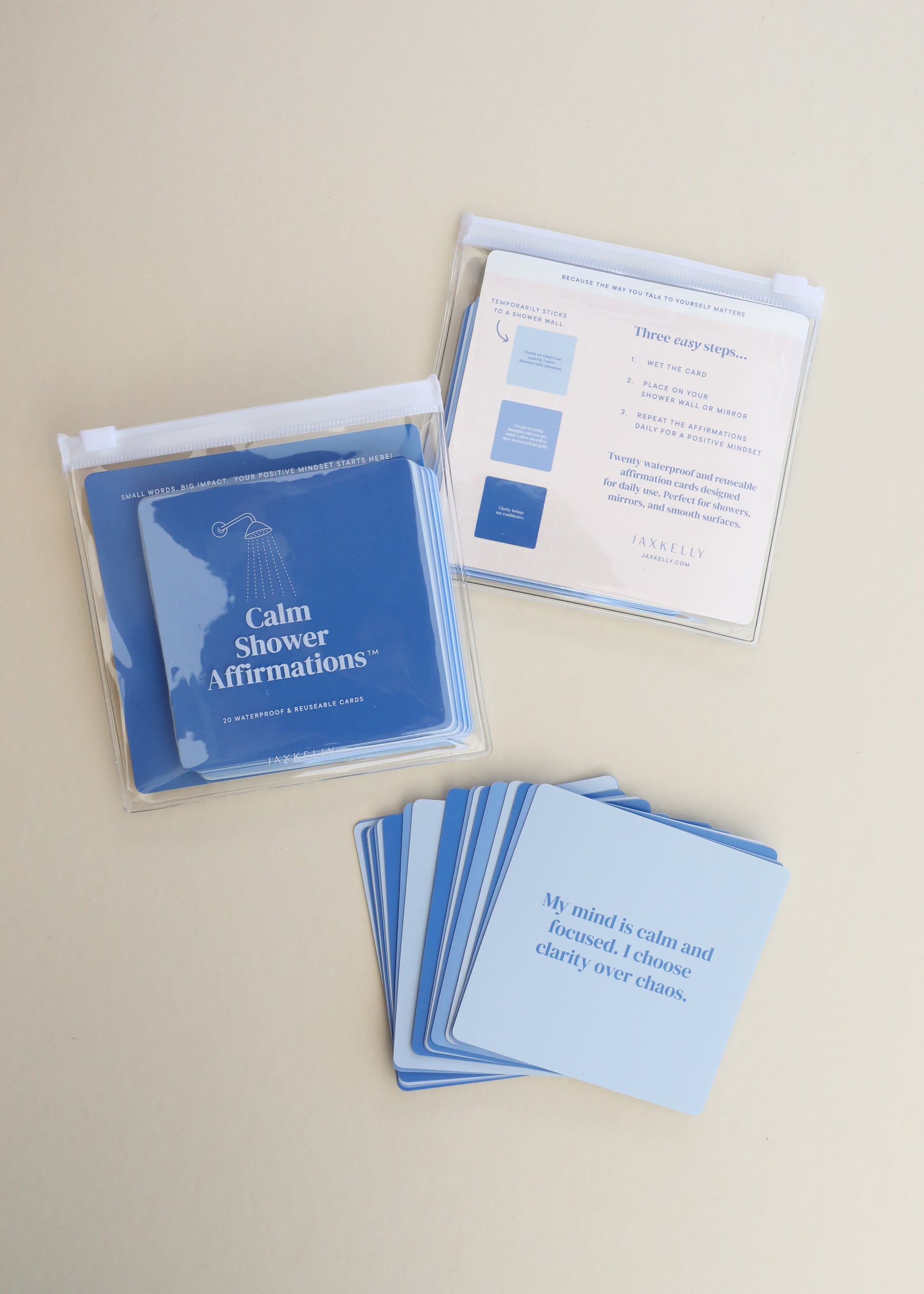 Calm Shower Affirmations™—a set of blue waterproof quote cards—is shown on a light surface. Some are spread out, others in a clear plastic case. One reusable card reads: My mind is calm and focused. I choose clarity over chaos.