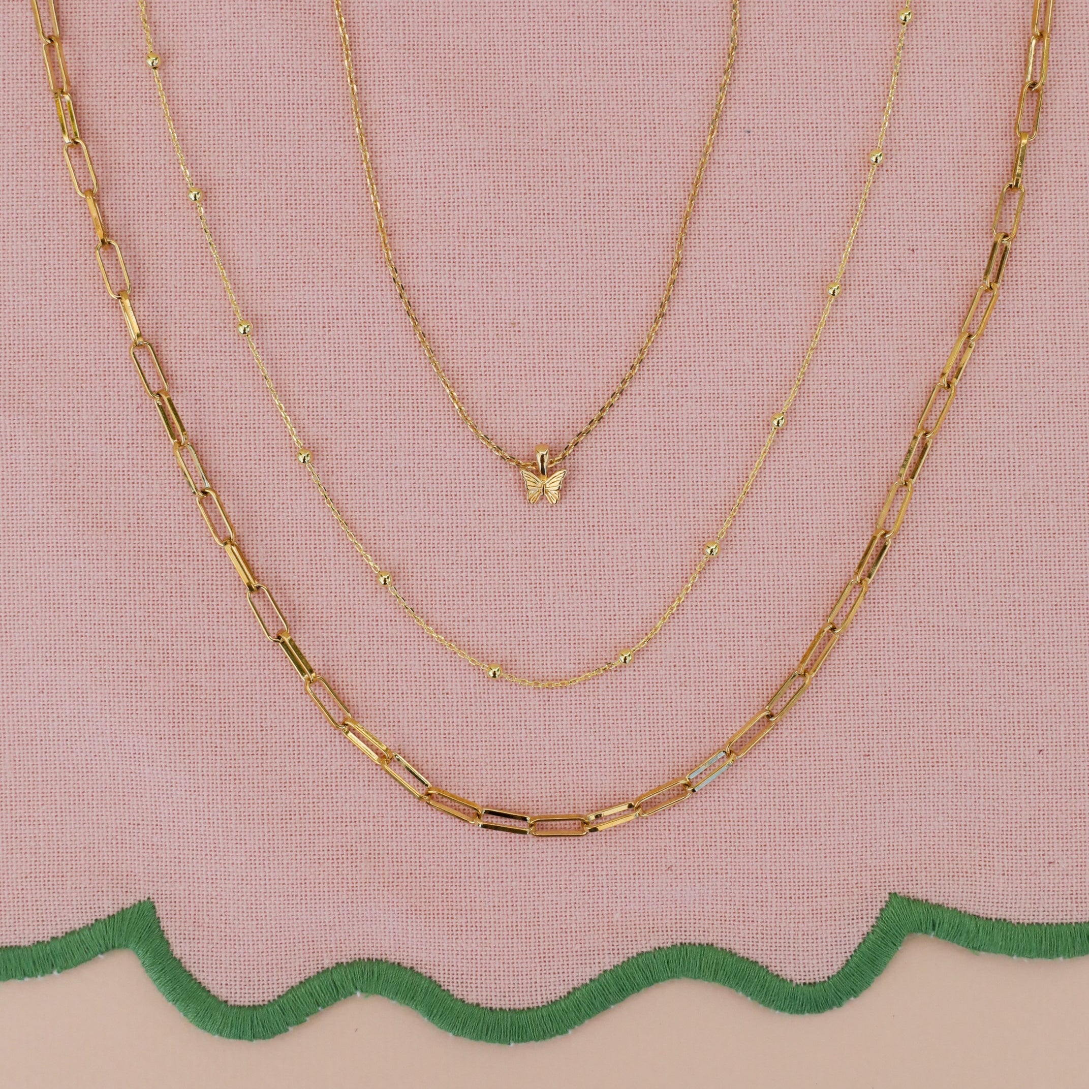Three gold JaxKelly necklaces displayed on pink fabric with a green scalloped edge: the Mystery Necklace has a butterfly pendant, another sparkles with hidden gems, and the third features elongated rectangular links.