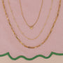 Three gold JaxKelly necklaces displayed on pink fabric with a green scalloped edge: the Mystery Necklace has a butterfly pendant, another sparkles with hidden gems, and the third features elongated rectangular links.