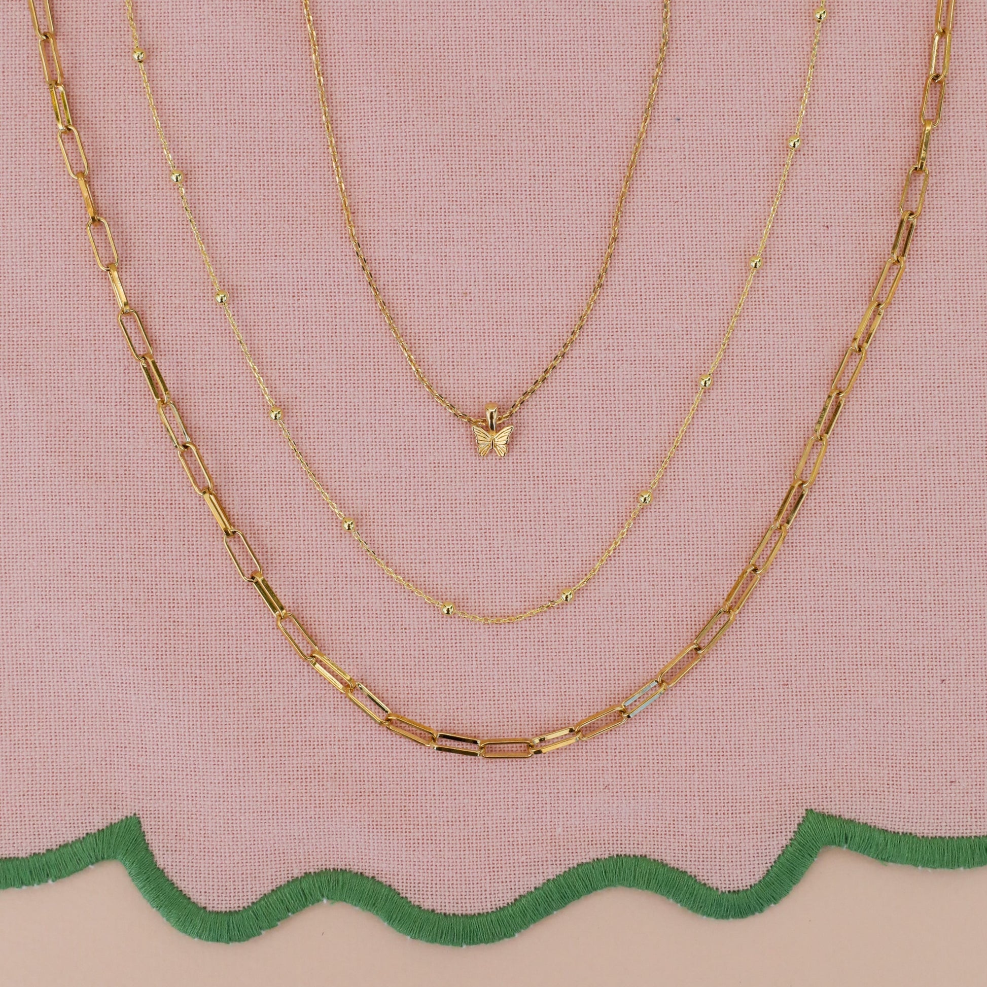 Three gold JaxKelly necklaces displayed on pink fabric with a green scalloped edge: the Mystery Necklace has a butterfly pendant, another sparkles with hidden gems, and the third features elongated rectangular links.