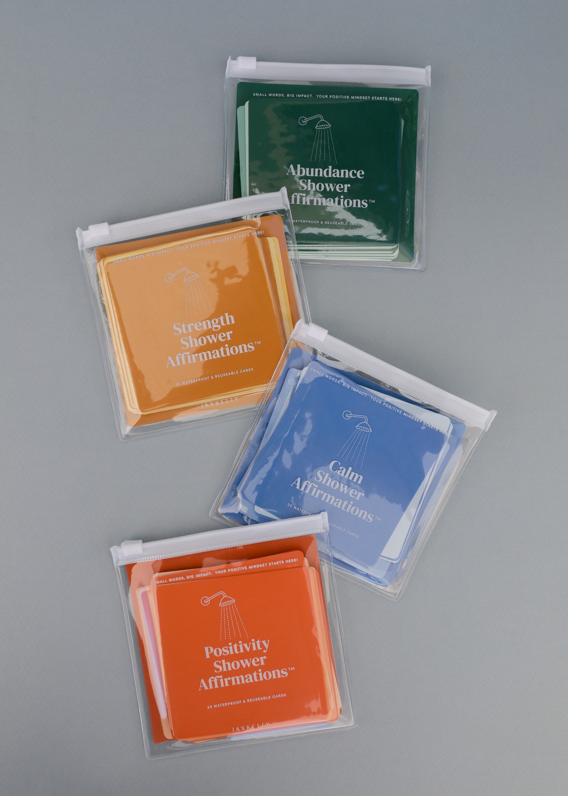 Four sets of colorful, waterproof Strength Shower Affirmations™ cards in clear pouches are arranged on a gray surface. The reusable quote cards are labeled: Abundance (green), Strength (orange), Calm (blue), and Positivity (yellow).
