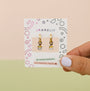 A hand with light purple nails holds a card featuring JaxKelly's Cupcake Enamel Charm Hoop earrings—gold hoops with cupcake charms—on a card decorated with colorful shapes and labeled hypoallergenic and Kid-Friendly Posts.
