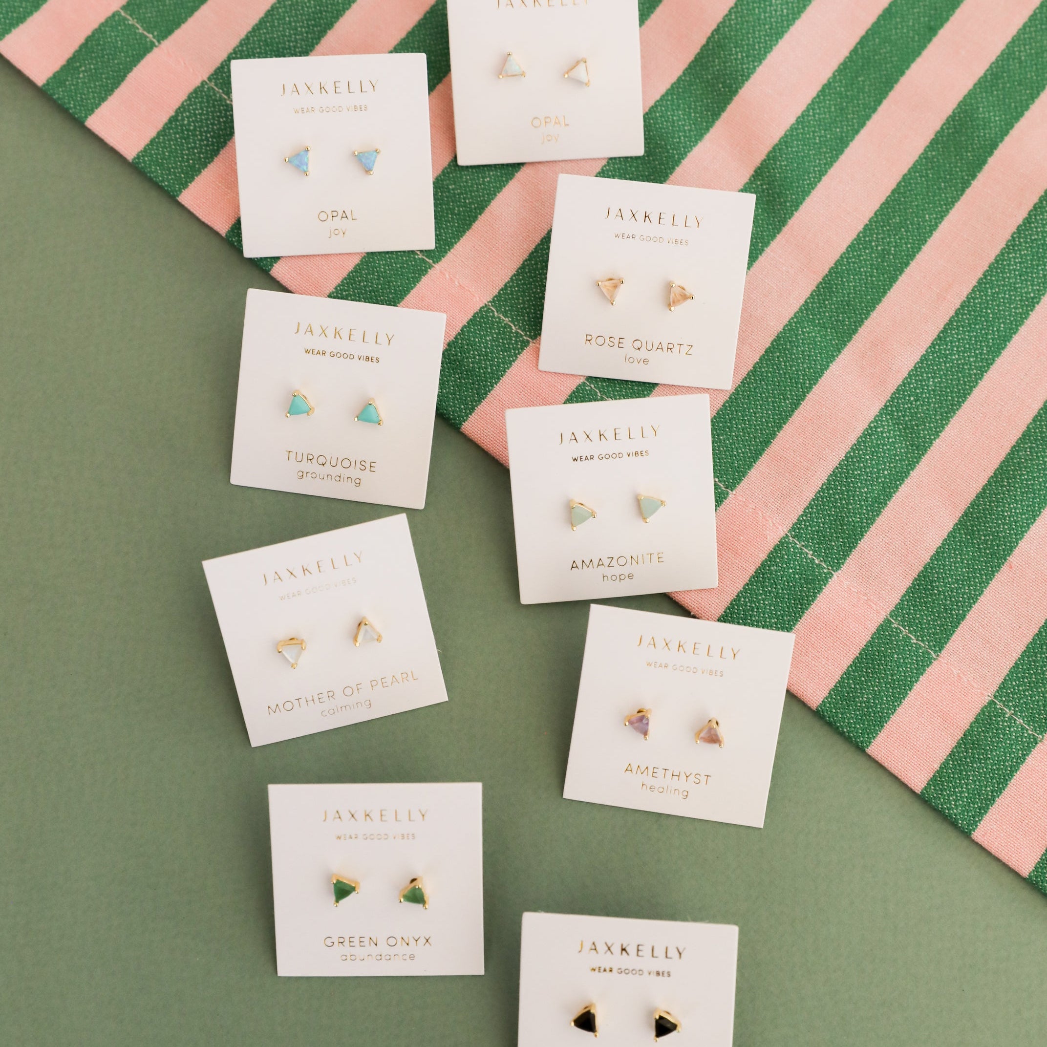 Eight pairs of JaxKelly Mystery Earrings on gemstone-labeled cards, arranged on green fabric beside a striped cloth—ideal for an earring sale or a surprise gift, featuring opal, turquoise, and rose quartz styles.