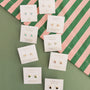 Eight pairs of JaxKelly Mystery Earrings, featuring surprise stones, are displayed on white cards over a green surface with a pink and green striped cloth in the corner.