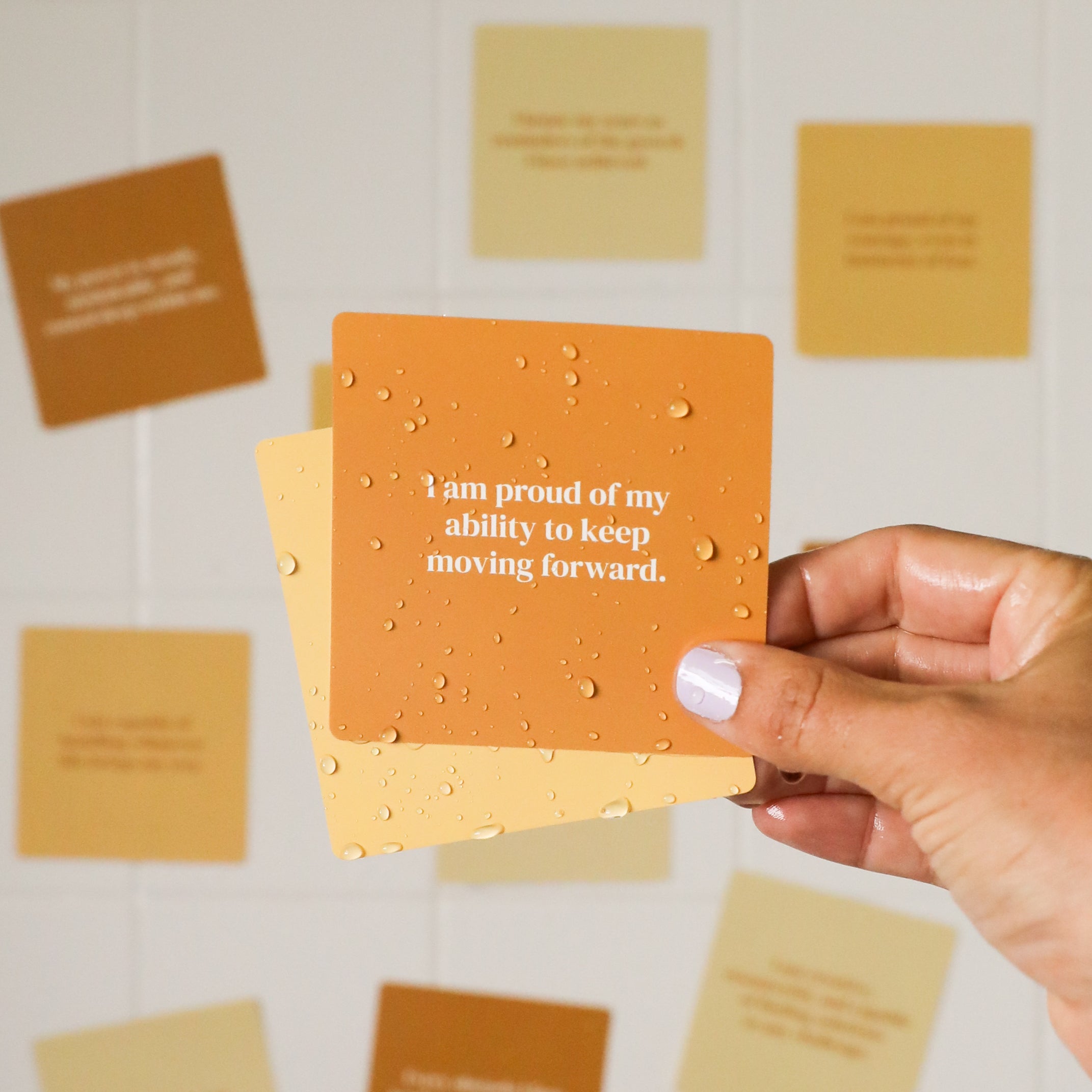 A hand with light purple nail polish holds a Strength Shower Affirmations™ orange waterproof card with water droplets, reading “I am proud of my ability to keep moving forward.” Blurred positive cards appear in the background on white tiles.