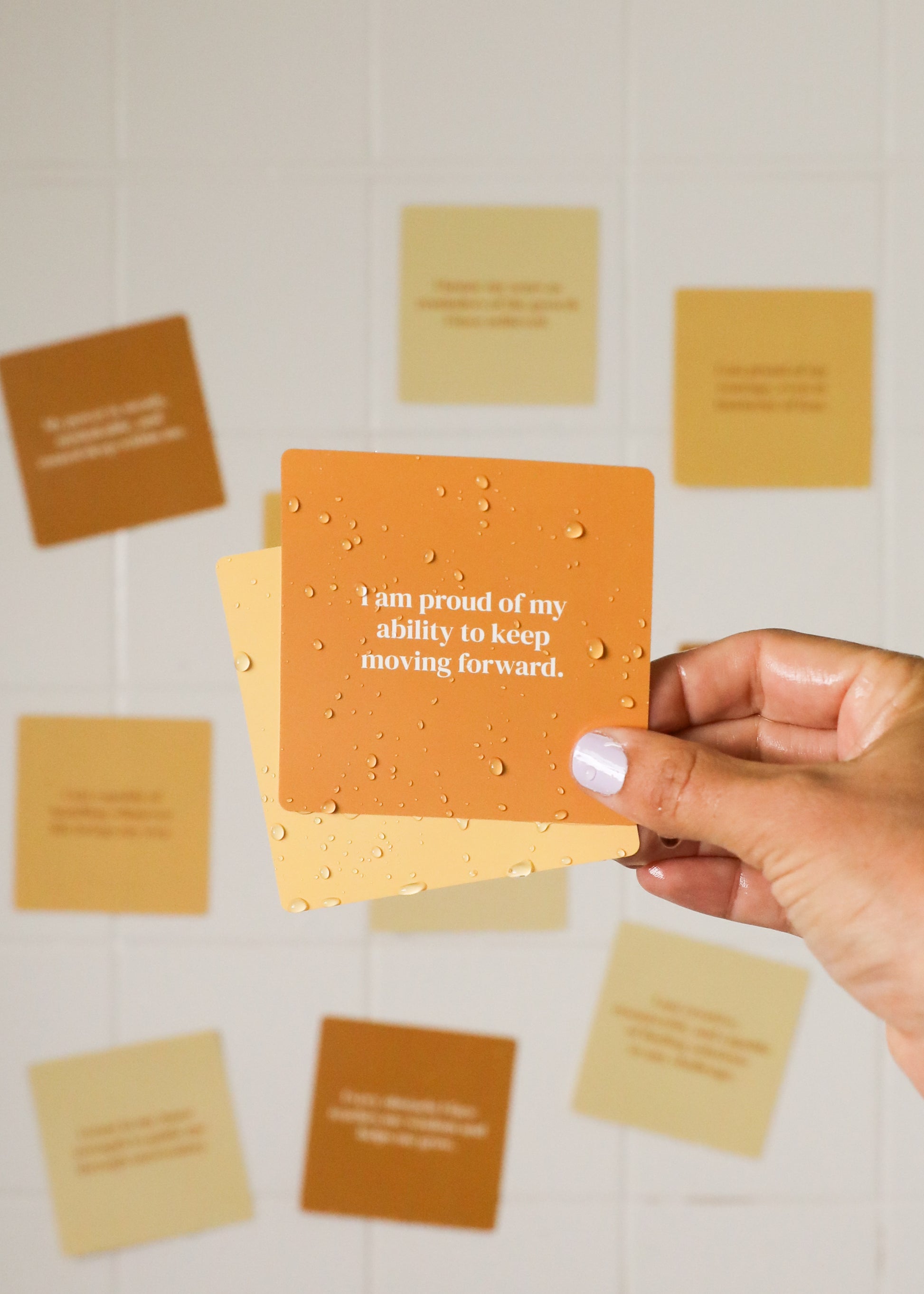 A hand with light purple nail polish holds a Strength Shower Affirmations™ orange waterproof card with water droplets, reading “I am proud of my ability to keep moving forward.” Blurred positive cards appear in the background on white tiles.