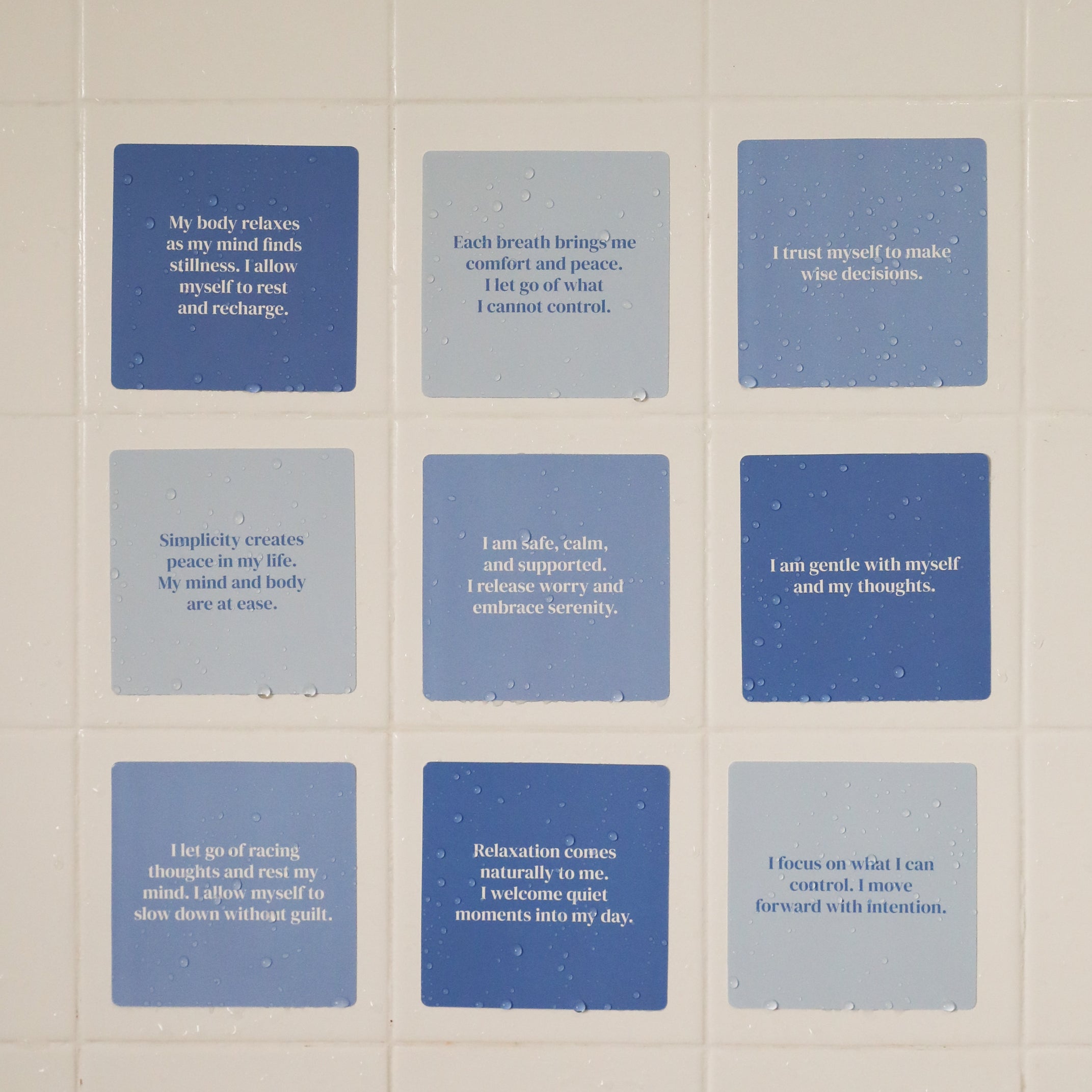 Nine Calm Shower Affirmations™ cards in blue and gray, each waterproof and printed with positive messages on relaxation, self-trust, and mindfulness, are displayed in a grid on a tiled white wall with visible water droplets.