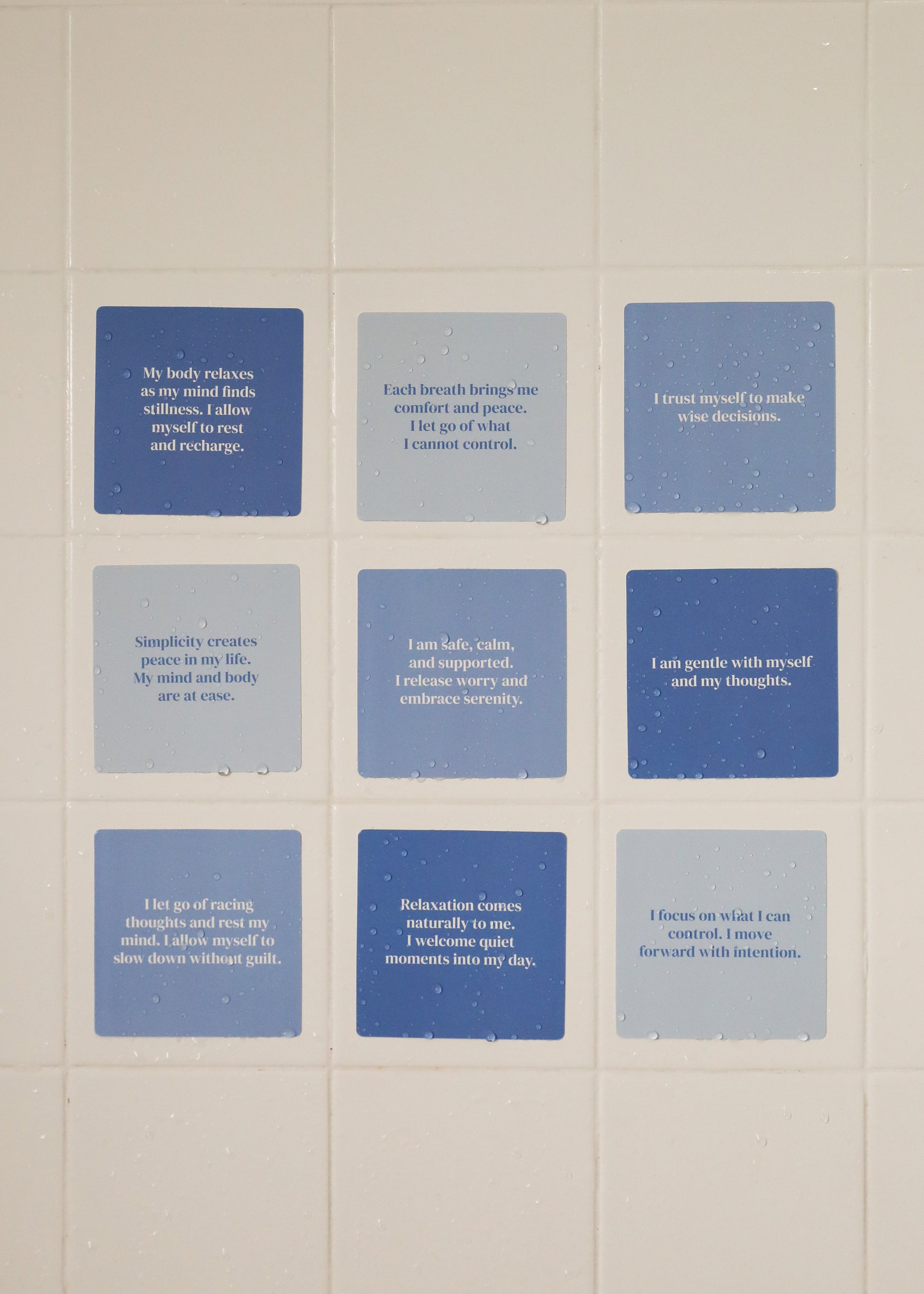 Nine Calm Shower Affirmations™ cards in blue and gray, each waterproof and printed with positive messages on relaxation, self-trust, and mindfulness, are displayed in a grid on a tiled white wall with visible water droplets.