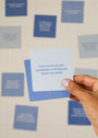 A hand with light nail polish holds a blue Calm Shower Affirmations™ card that reads, "I am centered and grounded. Each breath clears my mind," with other inspirational shower cards blurred in the background.