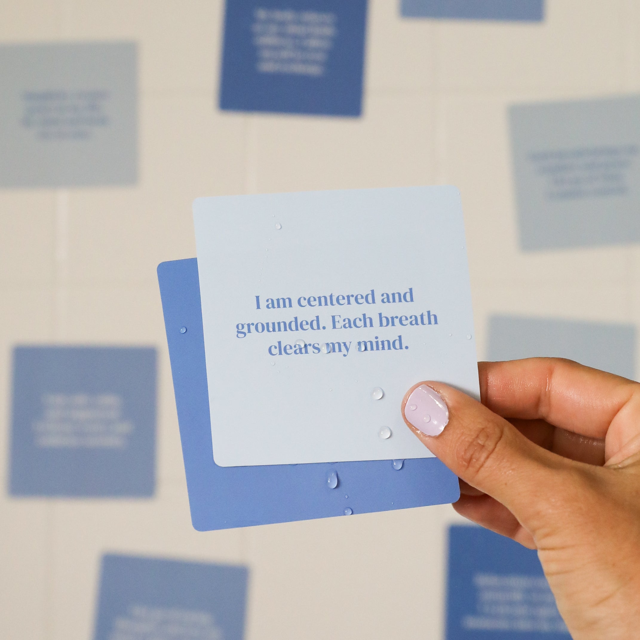 A hand with light nail polish holds a blue Calm Shower Affirmations™ card that reads, "I am centered and grounded. Each breath clears my mind," with other inspirational shower cards blurred in the background.
