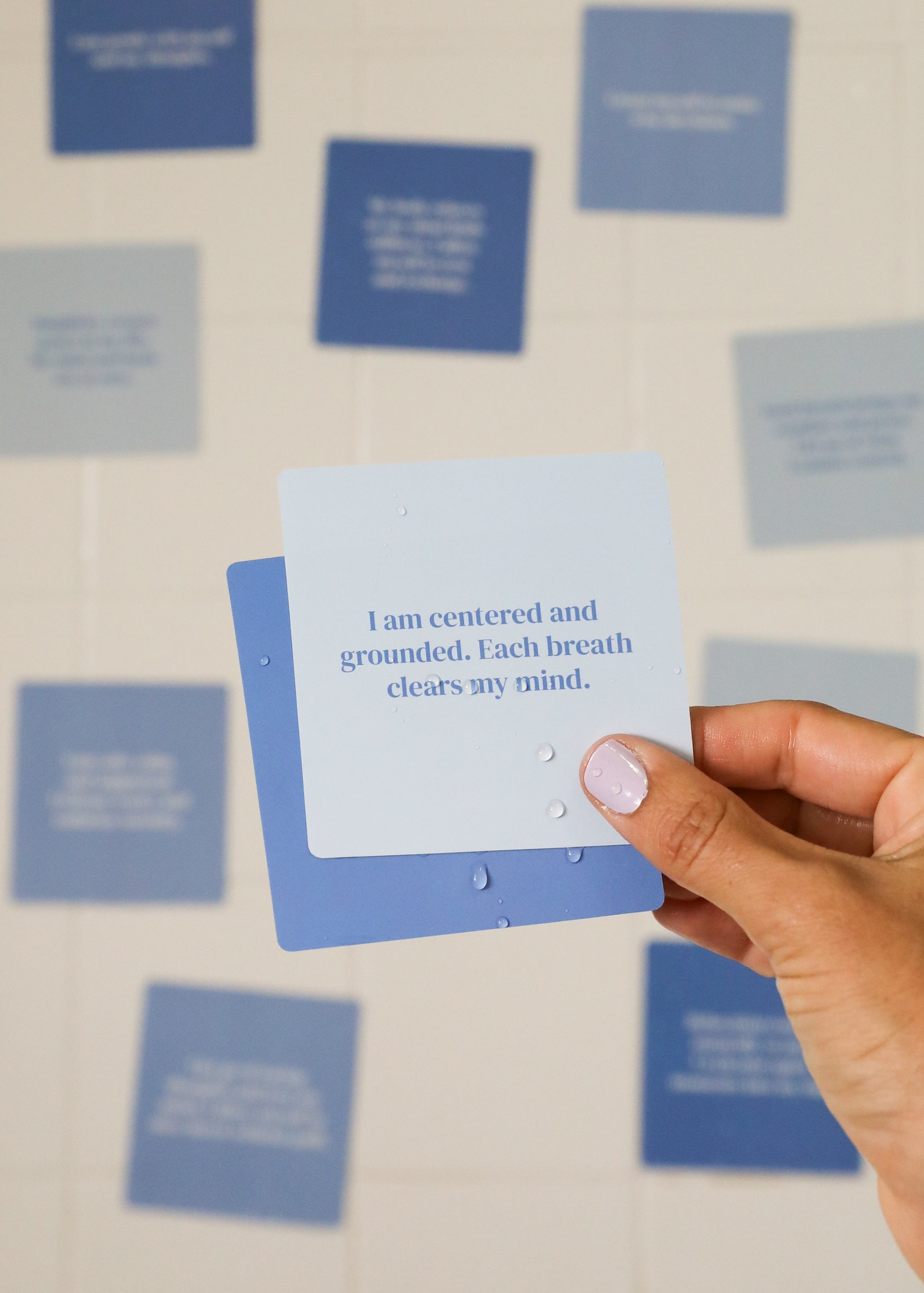 A hand with light nail polish holds a blue Calm Shower Affirmations™ card that reads, "I am centered and grounded. Each breath clears my mind," with other inspirational shower cards blurred in the background.