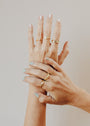 Two light-skinned hands with manicured pale and mint green nails are shown against a plain background, each adorned with stylish gold rings—featuring the Mystery Ring for a touch of intrigue.