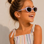 A smiling young girl wearing a pastel-striped dress and flower-studded sunglasses looks to the side, her hair in a bun. She accessorizes with JaxKelly’s Gumball Enamel Flat Back Kids Earrings—small, heart-shaped, hypoallergenic studs. Sunlight creates soft shadows against a light background.