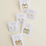 Five pairs of JaxKelly earrings, including Round - Amazonite Gemstone - Gold Earrings and other minimalist studs and drops in opal, rose quartz, and mother of pearl, are displayed on cards against a light cream background.