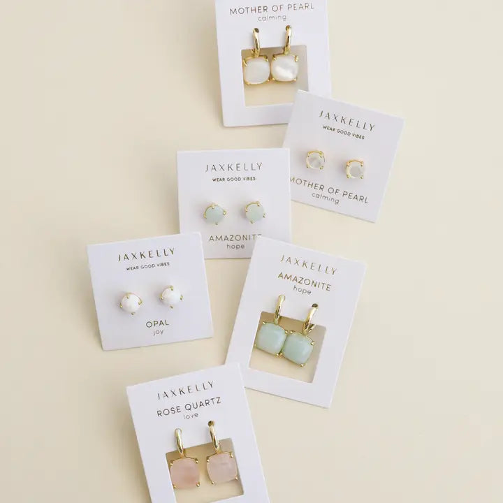Five pairs of JaxKelly earrings, including Round - Amazonite Gemstone - Gold Earrings and other minimalist studs and drops in opal, rose quartz, and mother of pearl, are displayed on cards against a light cream background.