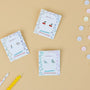 Three pairs of Artist Palette Enamel Flat Back Kids Earrings are displayed on decorative cards atop a beige surface, surrounded by pastel candies and two yellow candles. These hypoallergenic earrings are perfect for sensitive ears.