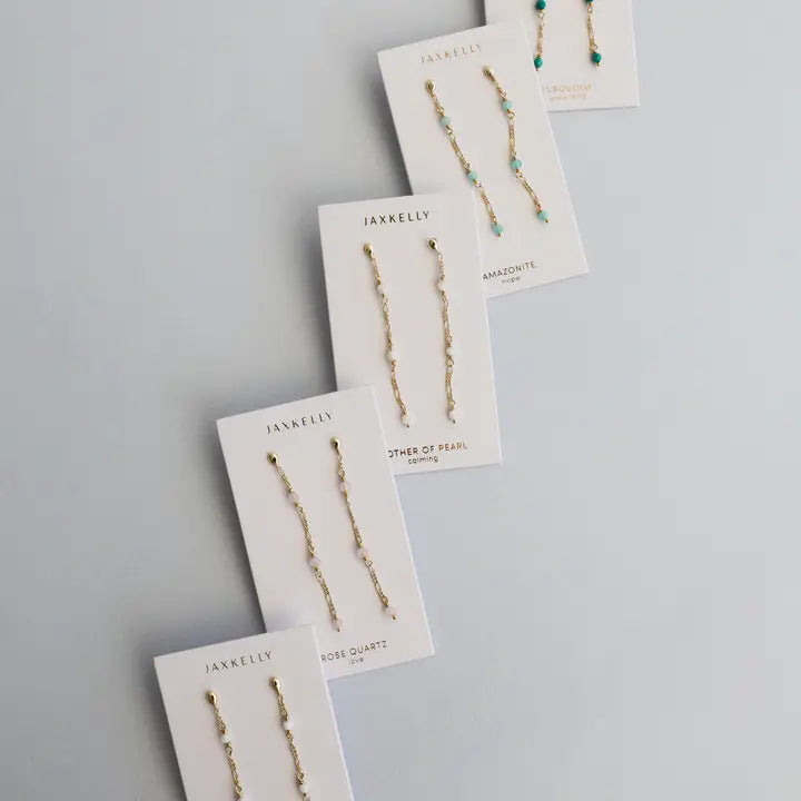 Five pairs of JaxKelly Gemstone Chain Drop earrings in Rose Quartz, featuring 18k gold plating and small gemstones, are displayed on white branded cards with gemstone names, arranged diagonally on a light gray background.