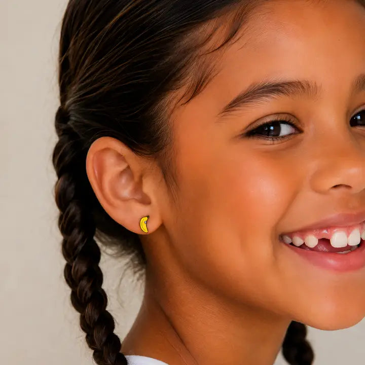 A smiling child with braided hair, wearing JaxKelly Banana Enamel Flat Back Kids Earrings, shows off a missing front tooth against a neutral background—perfect for kids with sensitive ears.
